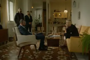 "Four people in a living room with mid-century decor; two seated and talking, two standing behind, surrounded by vintage furniture, patterned floor, plants, and warm lighting."